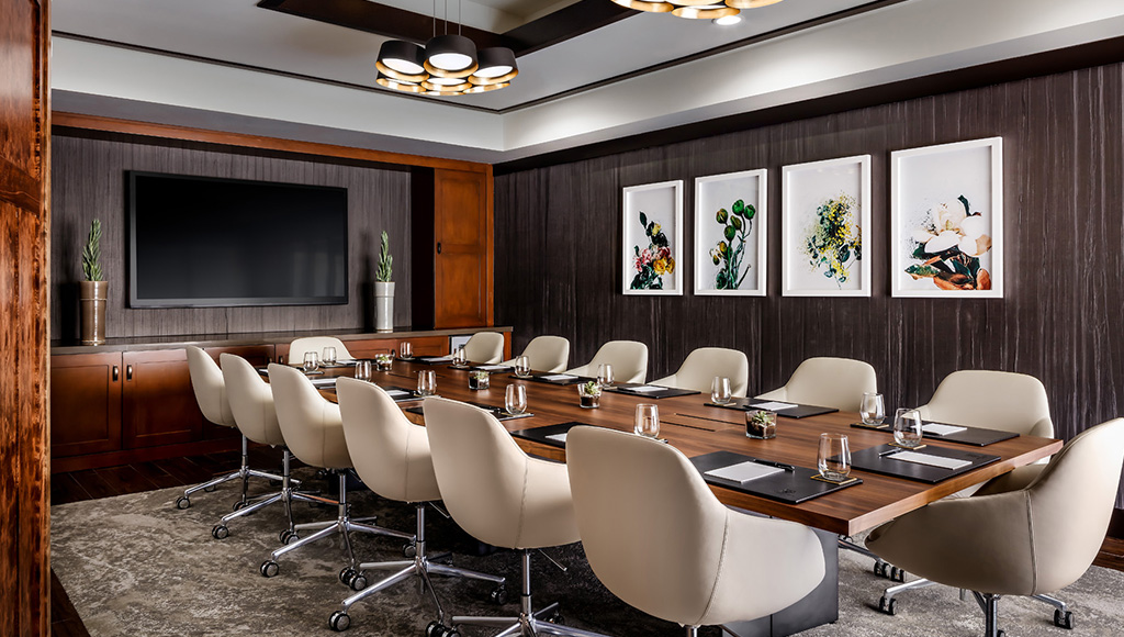 Meeting room with boardroom style seating arrangement; large TV along far wall, simple photos hanging on side wall; natural light coming in from windows