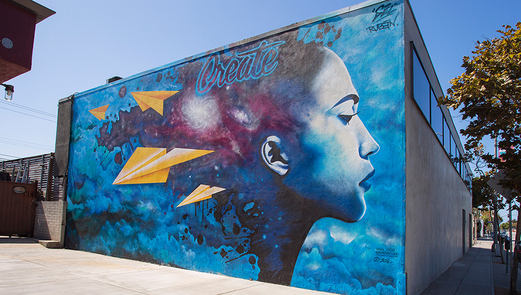 Blue mural of portrait of woman with paper airplanes coming out of her hair located on side of brick building