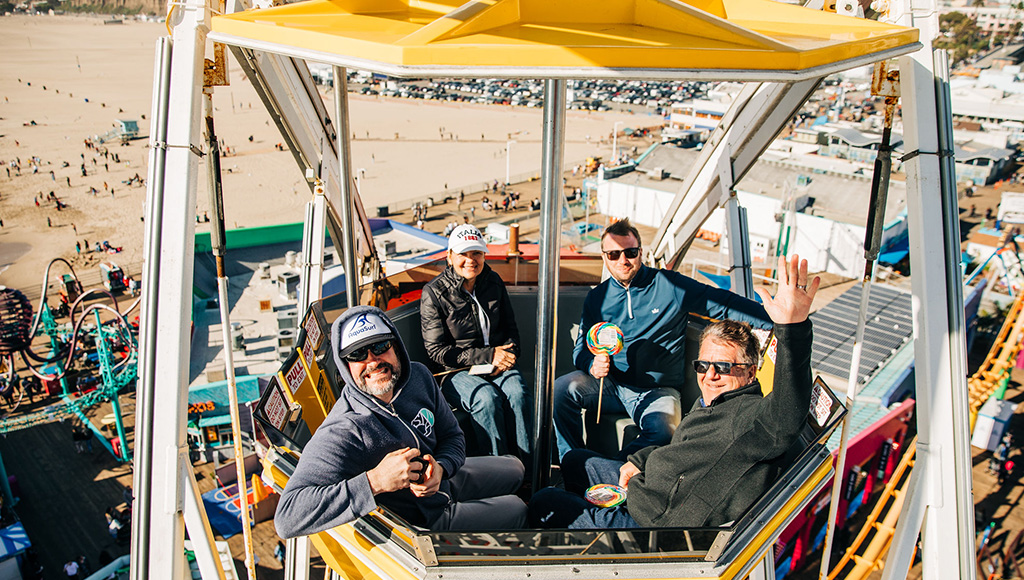 Four people sitting in yellow car at the top of the Ferris Wheel at Pacific Park, Santa Monica Pier; Santa Monica Beach in the background