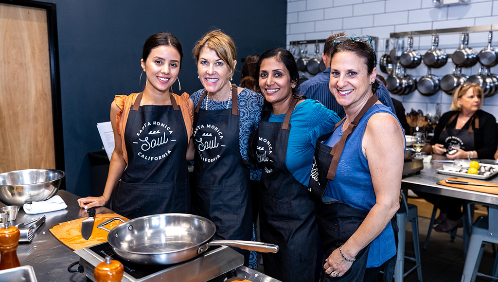 Four woman wearing black cooking aprons hugging and smiling at camera in a kitchen; pot on stove on the table in front of them