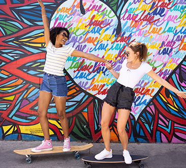 Two young women riding skateboards in front of colorful mural with multi-colored flowers and large heart with love written inside