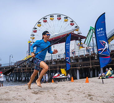 Man running on beach from the ocean with paddle; APP World Tour; Santa Monica Pier in background