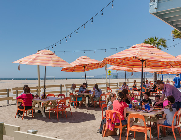 Outdoor dining in the sand at Santa Monica Beach; orange umbrellas providing shade to people sitting and eating