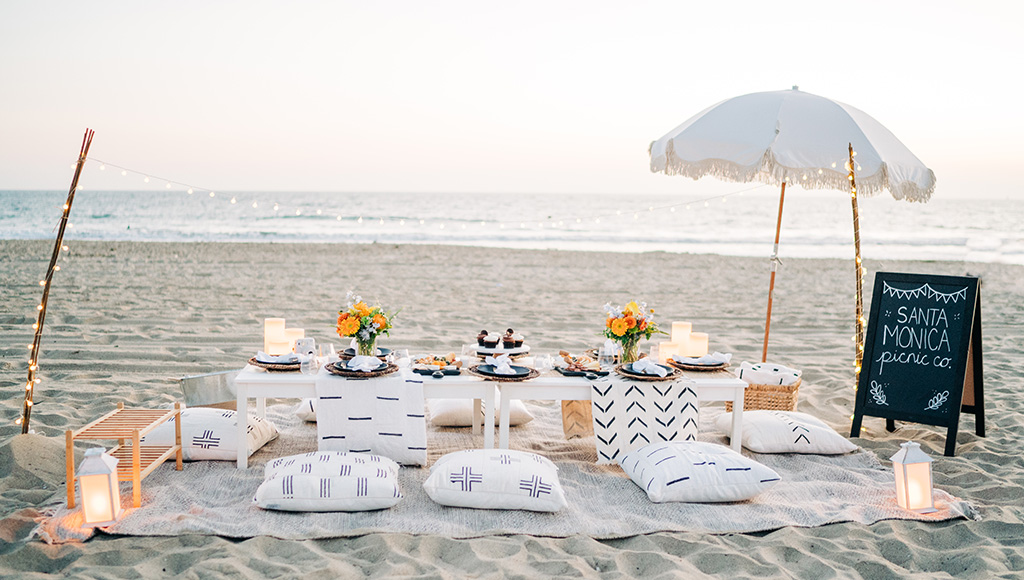 Picnic on Santa Monica Beach set up at sunset; low table with 8 pillows around it and food and candles set up; fairy lights strung on one side an an umbrella set up on the other