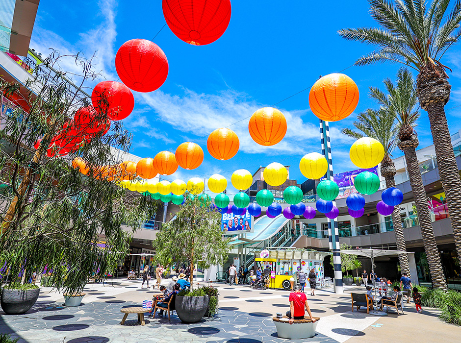 Rainbow chinese lanterns hanging over outdoor shopping plaza; people sitting and walking to shops