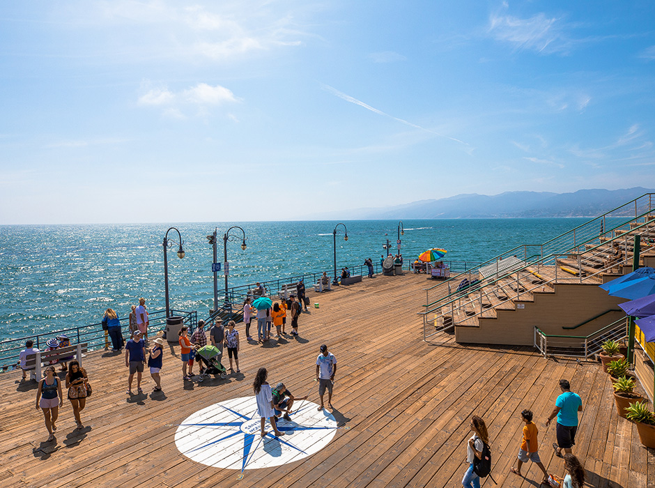 People walking and taking a photo at the end of Santa Monica Pier; End of pier overlooking ocean and hills in the distance