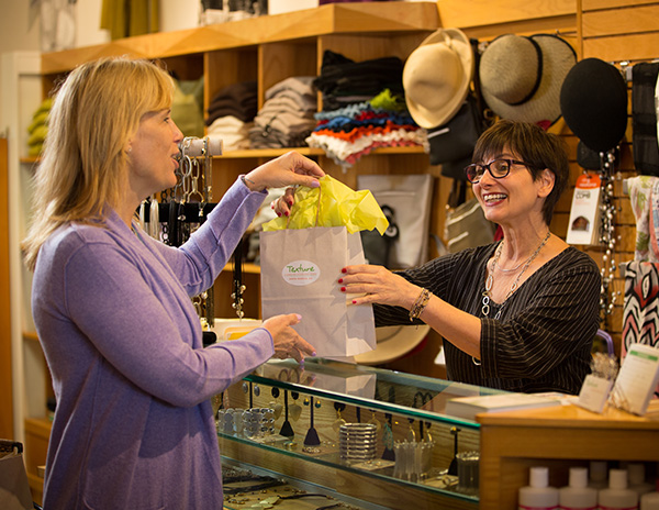 Cashier smiling and handing bag to customer; hats located on wall behind