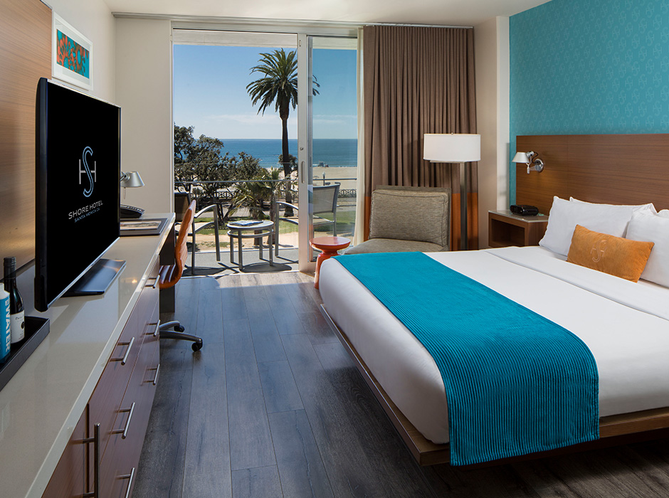 Hotel room with King bed on right-hand side of photo against wall. Large bed with blue throw, white sheets and an orange pillow; TV and counter to the right; open sliding door in front with curtains open and two chairs on outdoor patio overlooking Santa Monica Beach