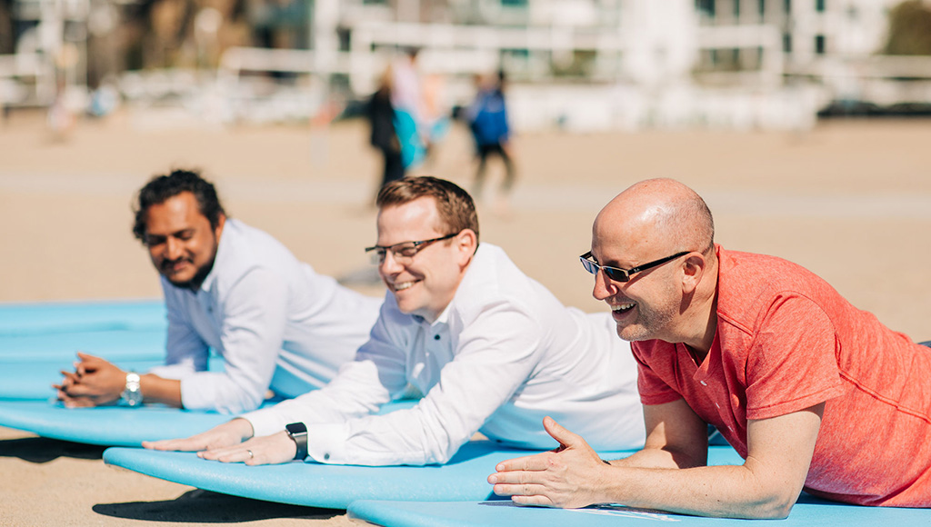 Three men in professional clothes smiling and laying on blue surf boards in the sand at Santa Monica Beach