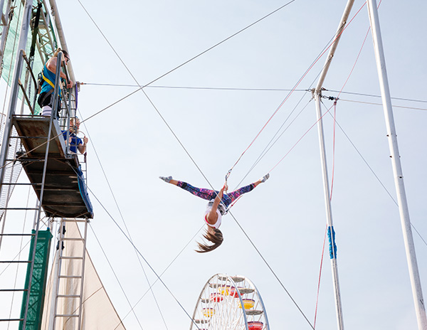 Woman upside down doing trapeze flip; Santa Monica Pier's Pacific Wheel in background