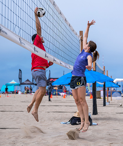 Man and woman jumping up across volleyball net from each other; man spiking ball; woman and man playing beach volleyball