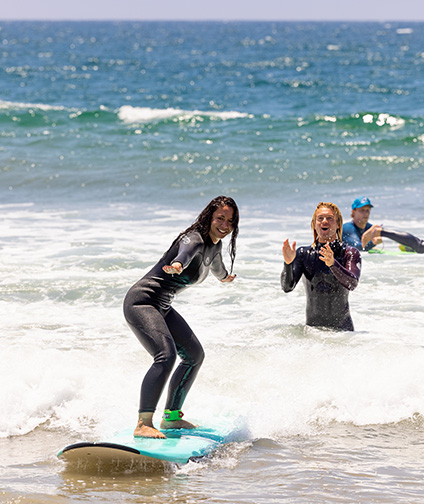 Woman standing on surfboard in ocean; teacher cheering behind her