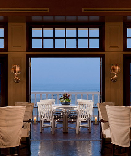 Looking down hotel dining room to open double doors; two plush chairs set up on outdoor deck with table and roses; beach in the background; sunset dinner