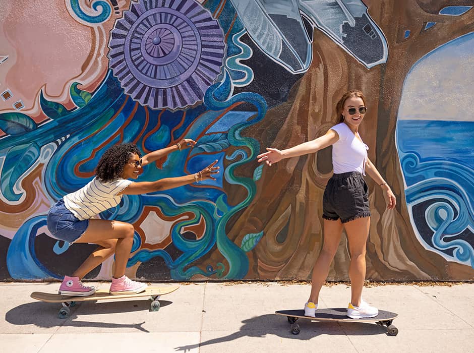 Photo of two girls on skateboards in front of mural; girl in front reaching behind to hold hands with girl skating behind
