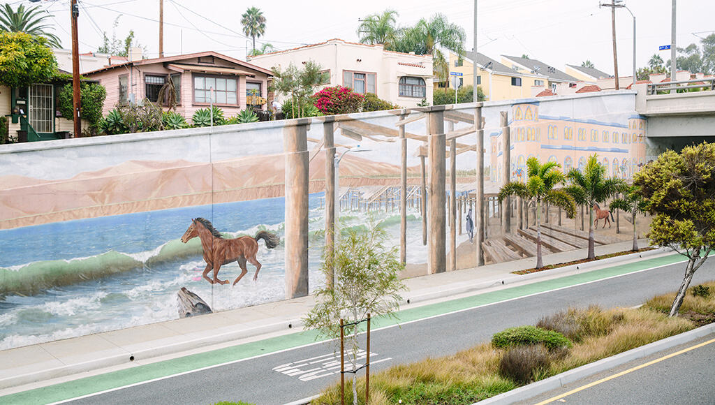 Picture taken across street from long wall leading to underpass with mural of horses running freely along beach painted